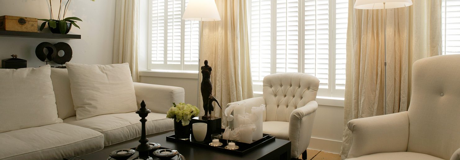 White window shutters on three windows in a living room with cream chairs.